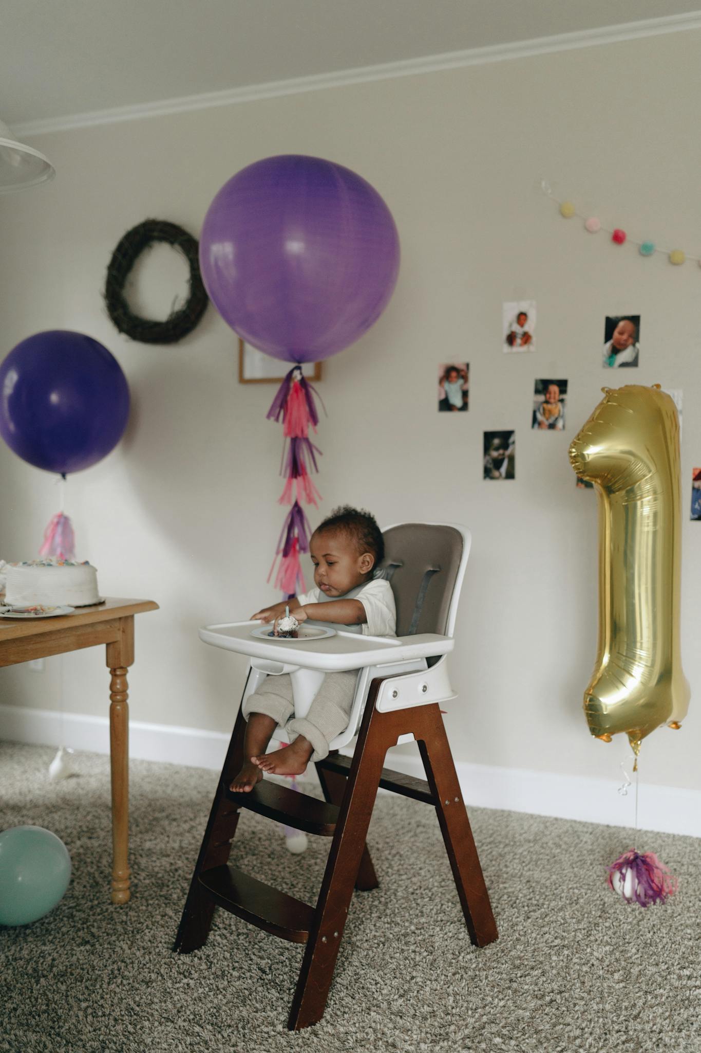 Adorable baby in high chair celebrating first birthday with balloons and cake.